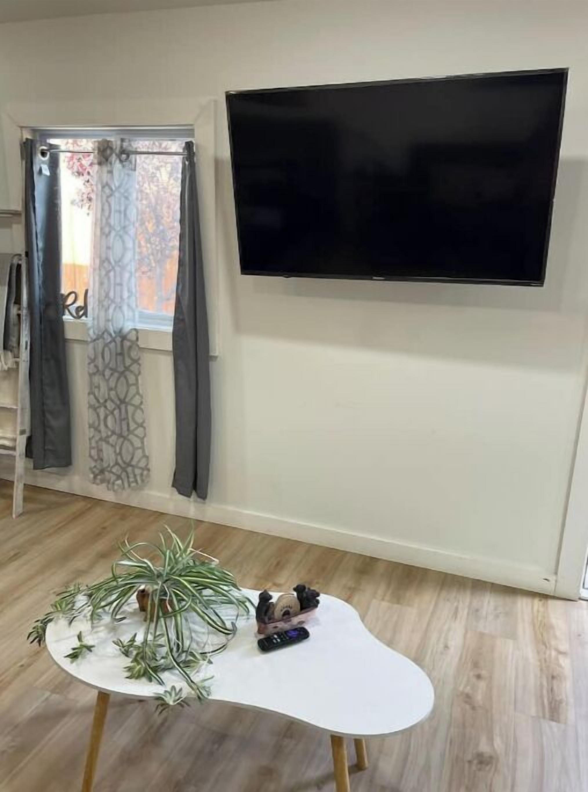 Une pièce dotée d’un téléviseur à écran plat fixé au mur, d’une table blanche ornée d’une plante et de télécommandes, et d’une fenêtre avec des rideaux à motifs.