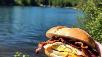 Une main tenant un hamburger garni de bacon et de fromage, avec un lac et de la verdure en arrière-plan.