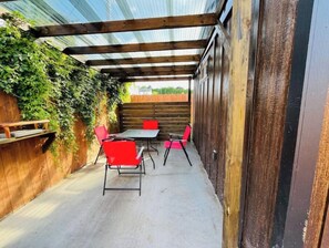 Une terrasse extérieure avec une table et quatre chaises rouges, entourée de murs en bois et surmontée d’un toit en métal.