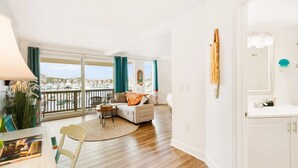 A bright living room with a balcony overlooking a marina, a white sofa with cushions, a small round table, and a view of the marina.