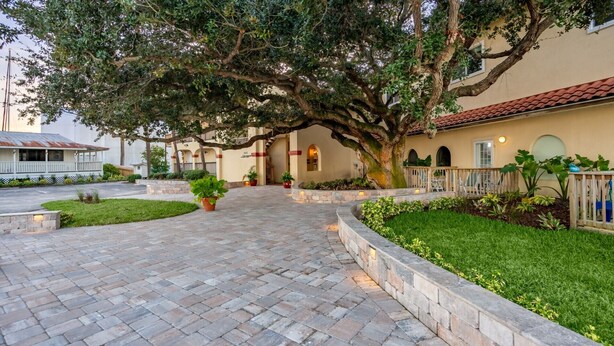 A paved pathway leading to a building with a large tree, a well-maintained lawn, and a fence with blue chairs. - History Meet Beach! King Studio, Sunroom and Balcony (St. Augustine)