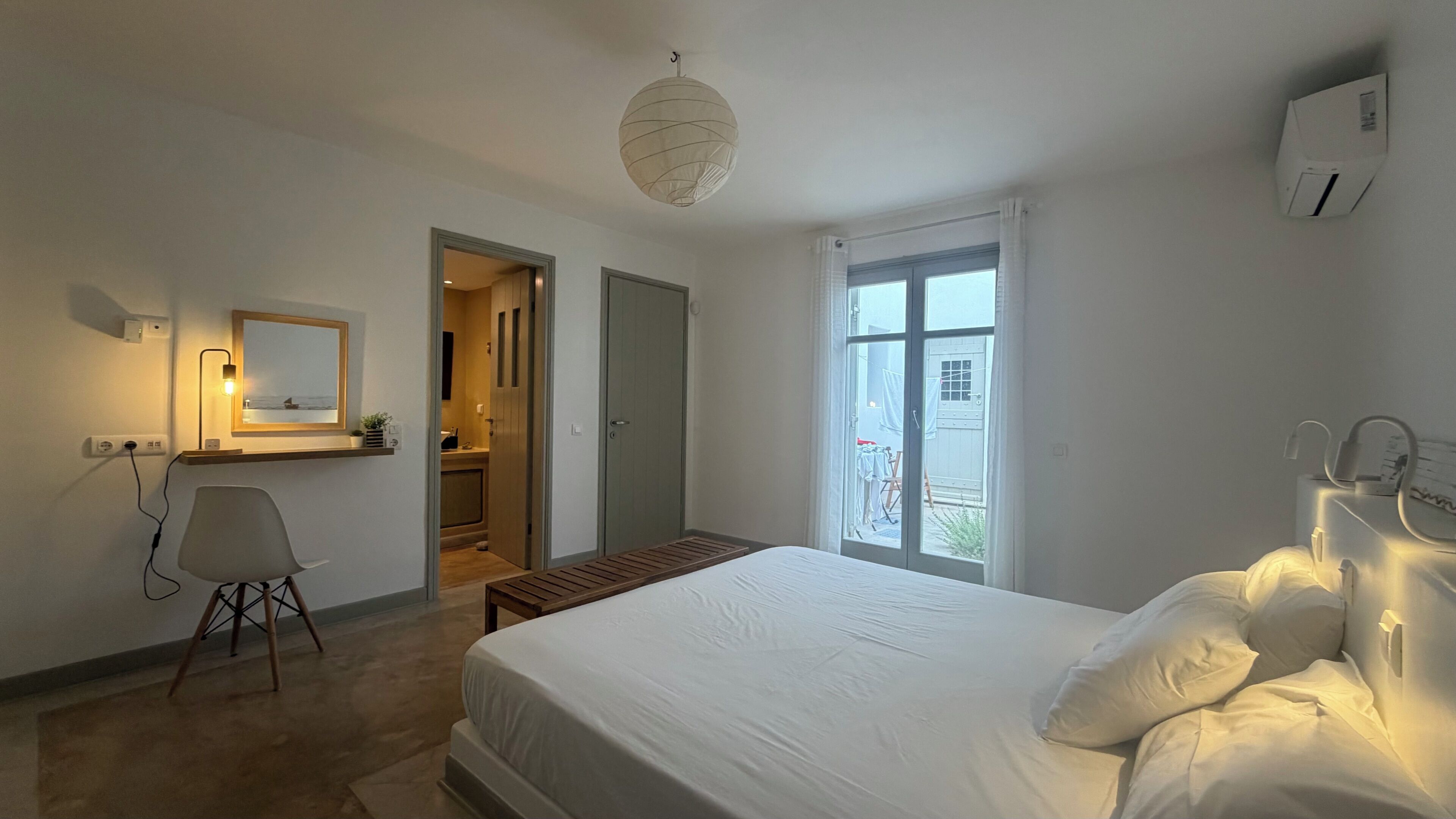 A bedroom with a bed, bedside table, lamp, and a view of the outside.