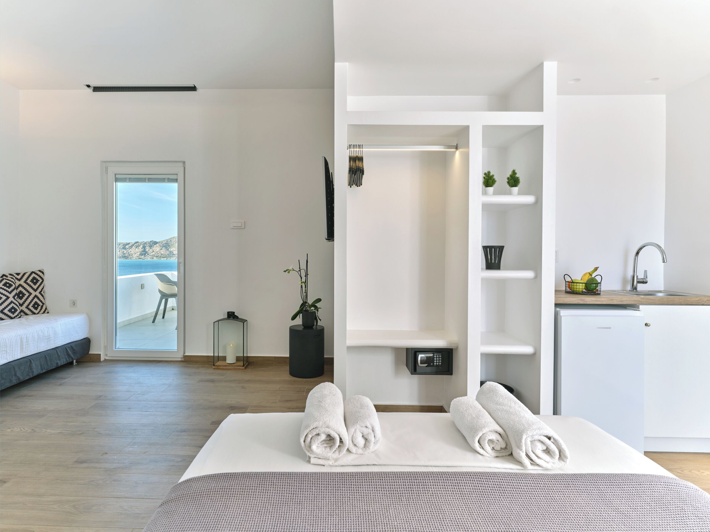 A modern hotel room with a bed, a view of the sea, a small balcony, a kitchenette, and a bathroom.