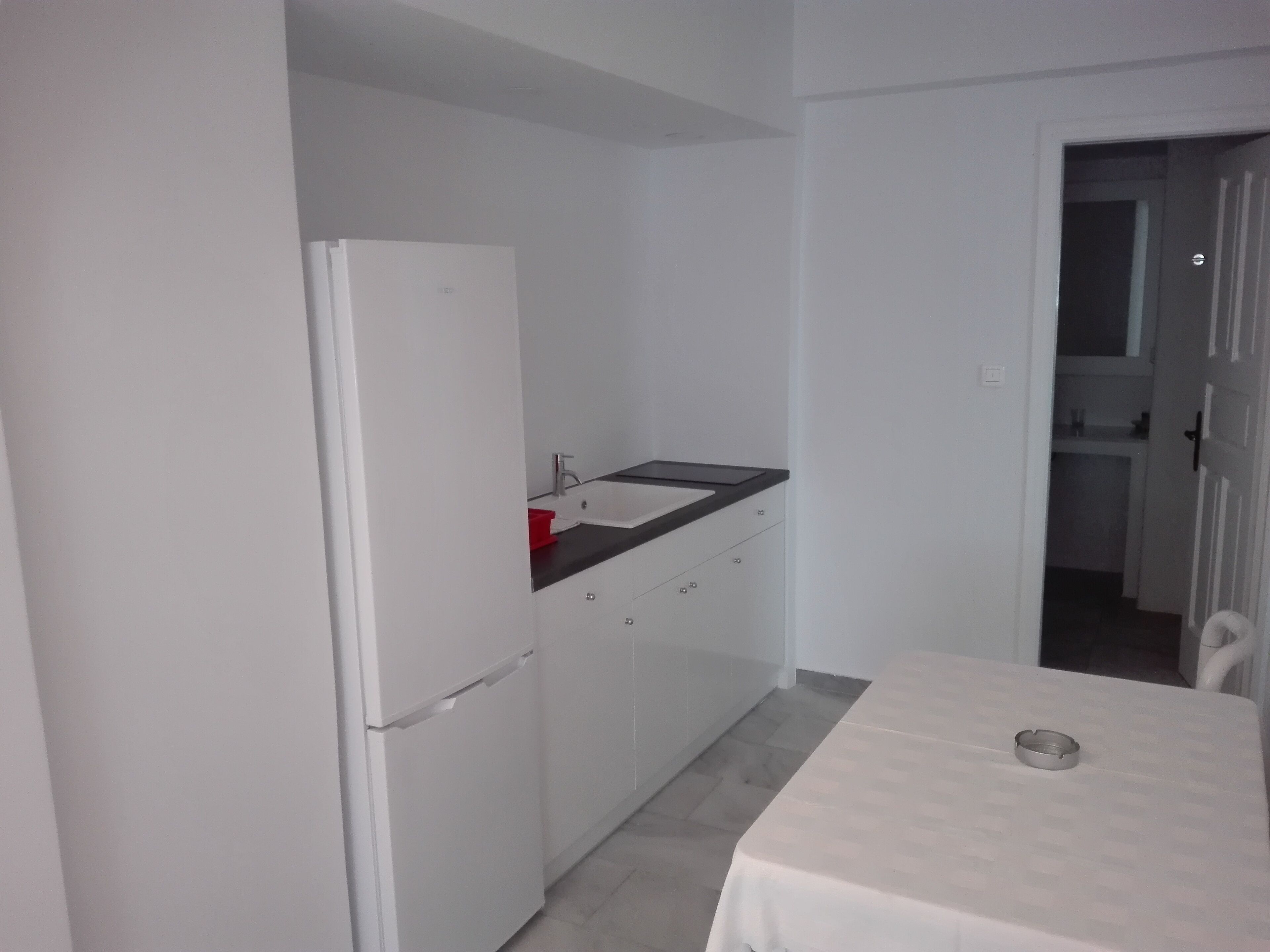 Uma cozinha compacta com armários brancos, uma geladeira branca, bancada preta e uma pequena mesa com bandeja de metal.