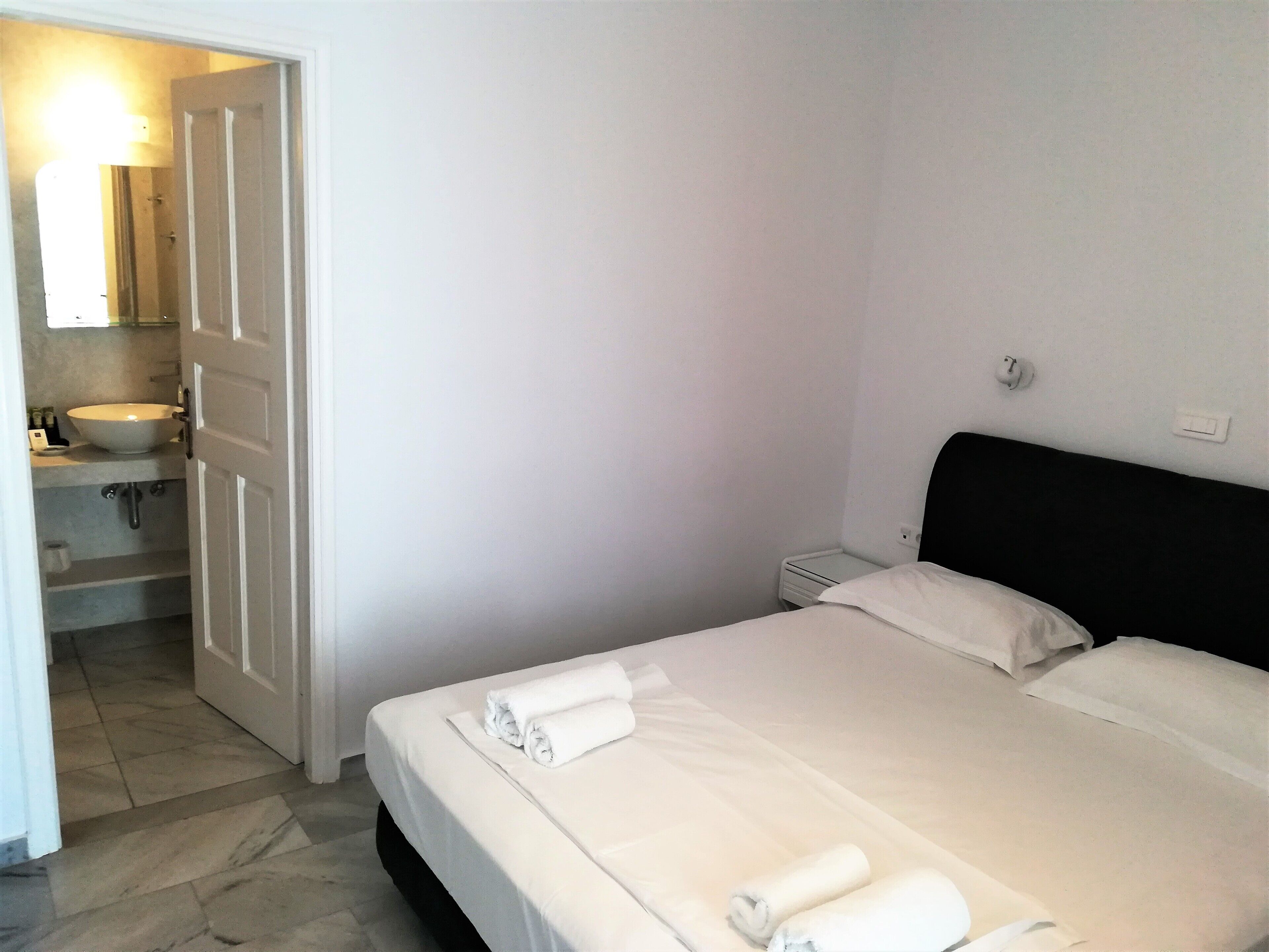 Une chambre d’hôtel comprenant un lit, une table de chevet, une commode, une salle de bain avec un lavabo et un miroir, et une porte donnant sur une autre pièce.