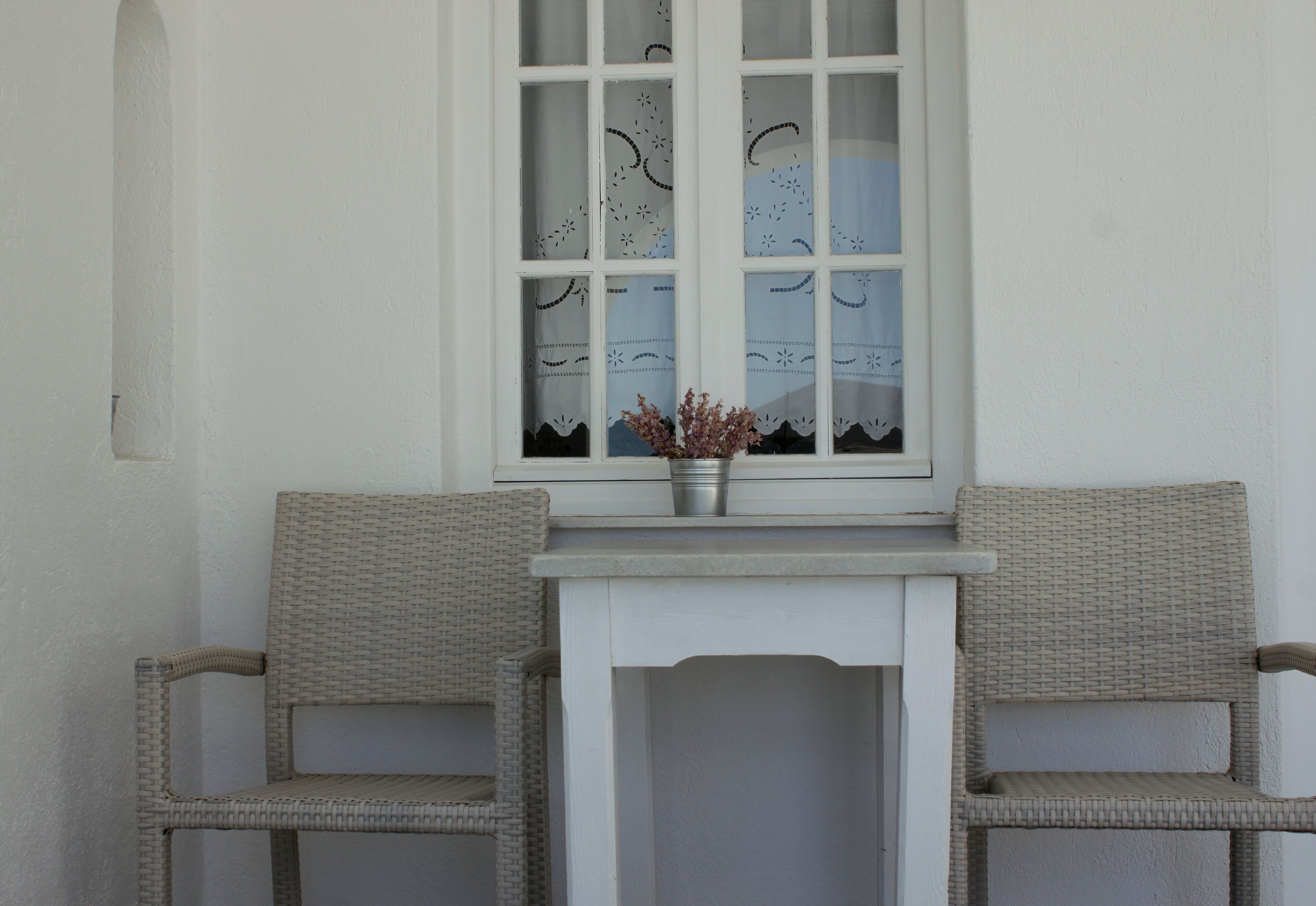 A white-framed window with patterned curtains, a small table with a potted plant, and two wicker chairs.