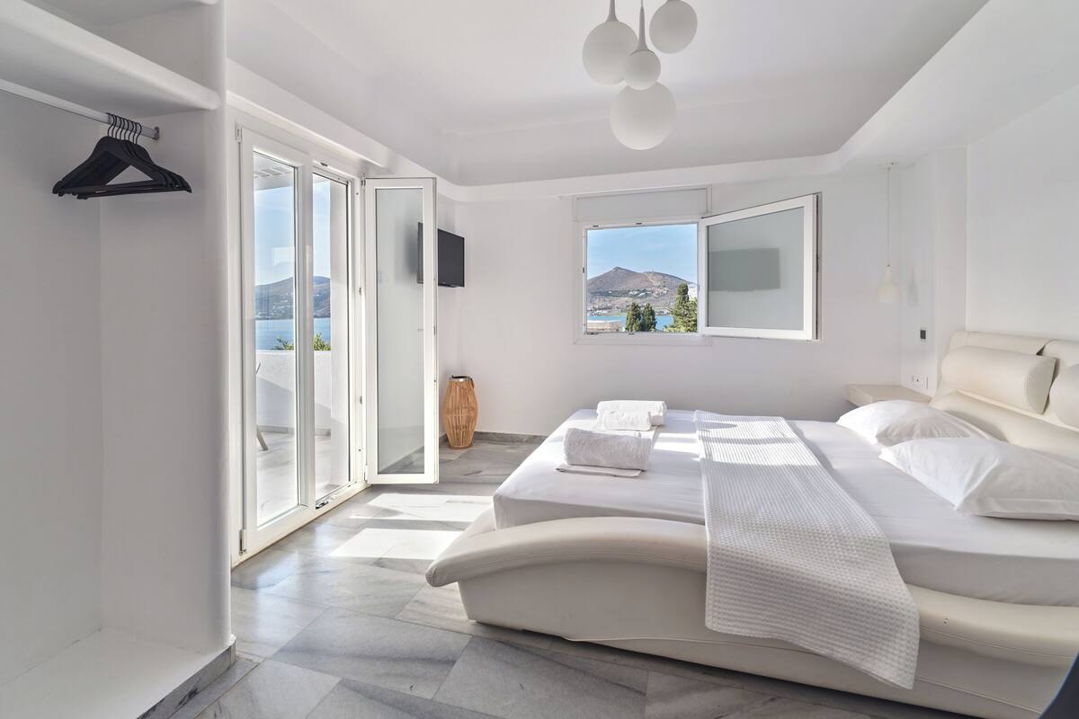 A modern bedroom with a large bed, a view of the sea, and a sliding glass door leading to an outdoor area.