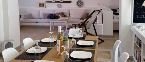 Un comedor con una mesa de madera dispuesta para seis comensales, sillas blancas, una araña y una zona de estar acogedora al fondo.