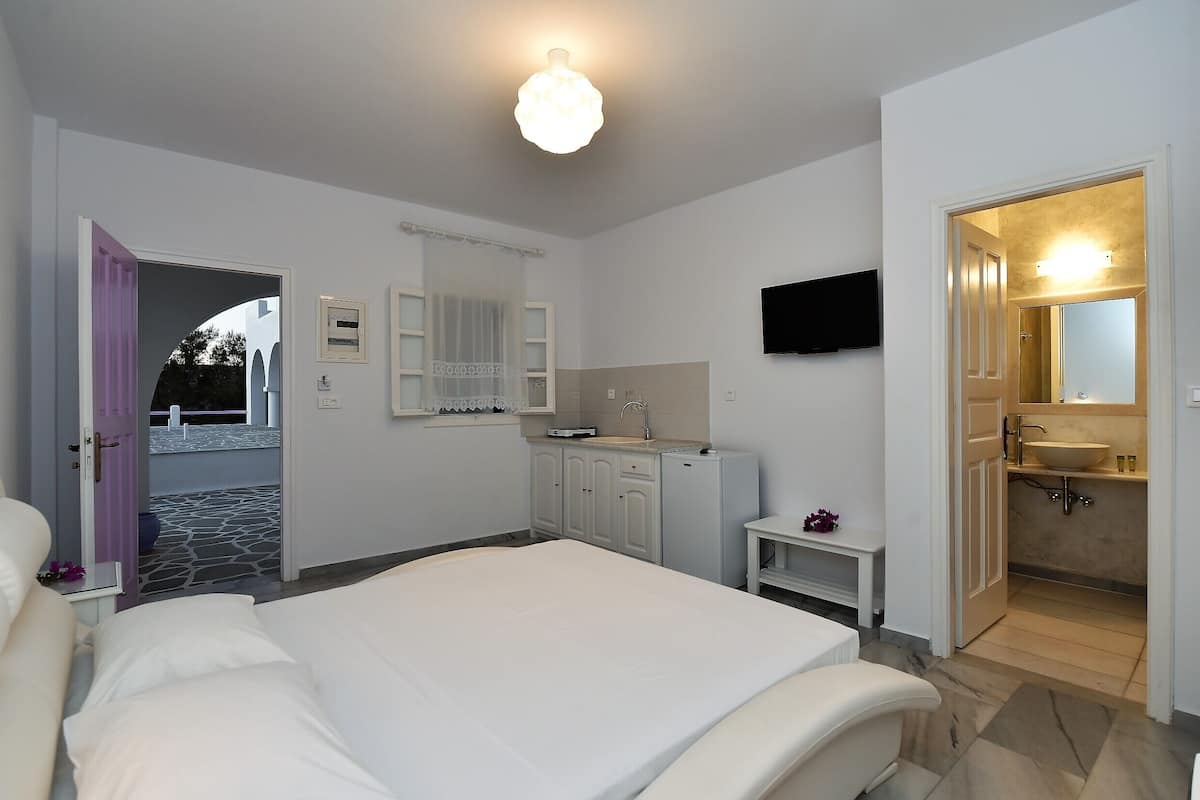 A bedroom with a bed, a small bedside table, a mirror, a TV mounted on the wall, and a bathroom with a sink and mirror.