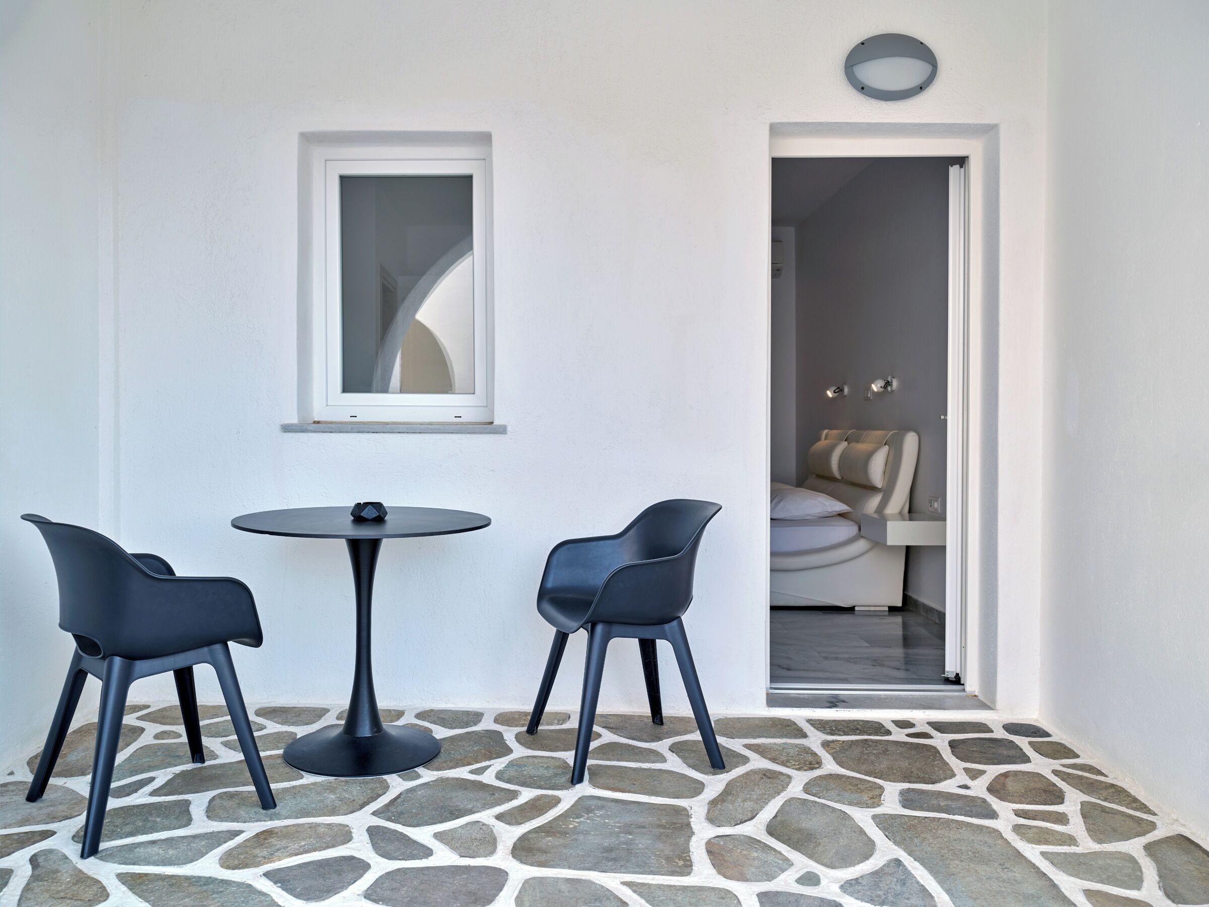Una habitación pequeña con suelo de piedra, una mesa redonda y dos sillas, una ventana y un acceso a otra habitación con un sillón de masaje.