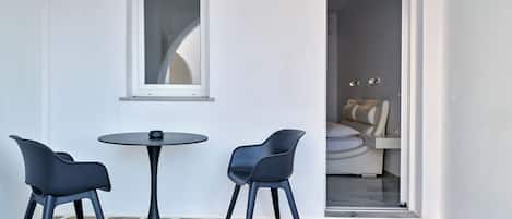 A small, stone-tiled room with a round table and two chairs, a window, and a doorway leading to another room with a massage chair.