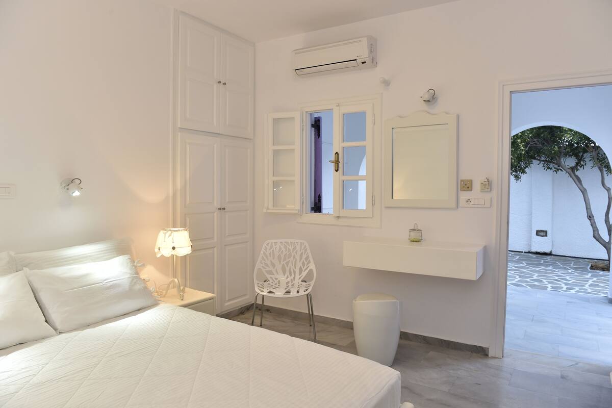 A white bedroom with a bed, a nightstand, a chair, a vanity with a mirror, and an air conditioning unit.
