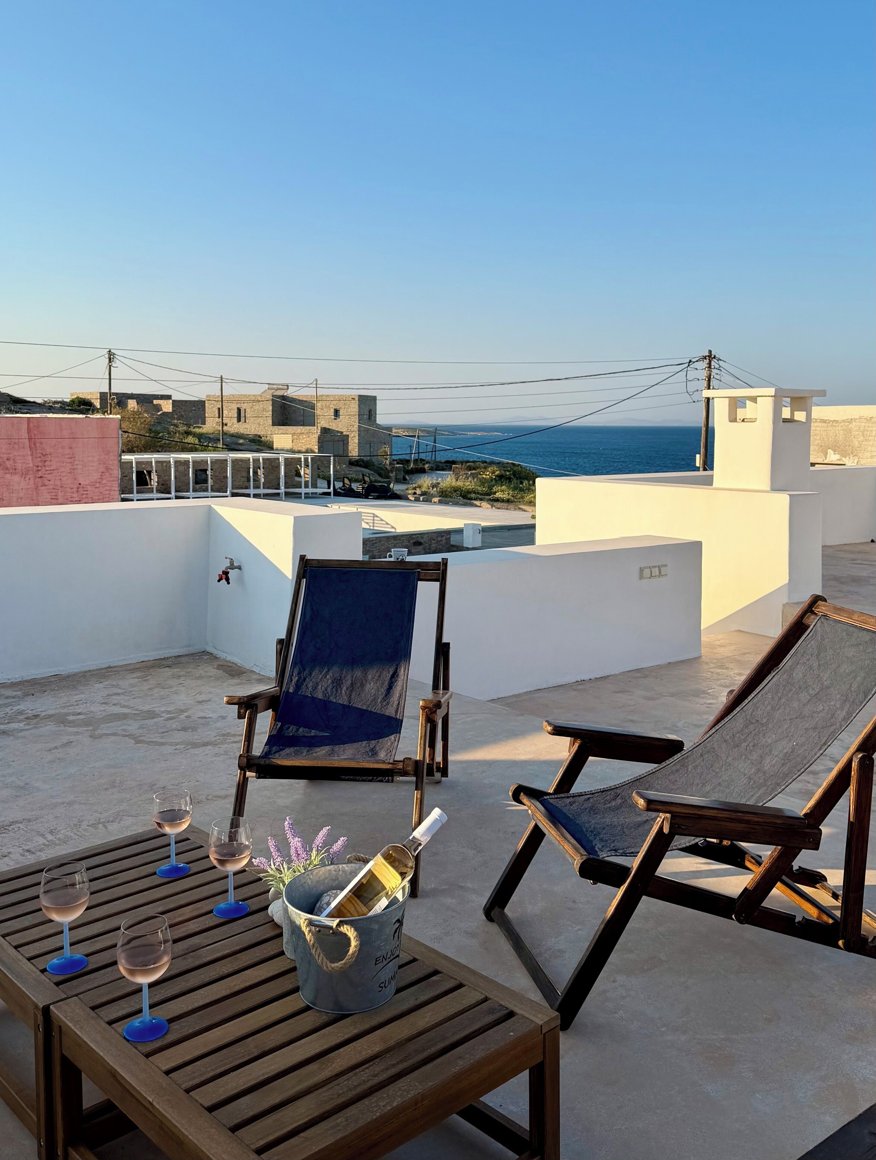 A rooftop terrace with two lounge chairs, a table with wine glasses and a bottle, and a view of the sea.