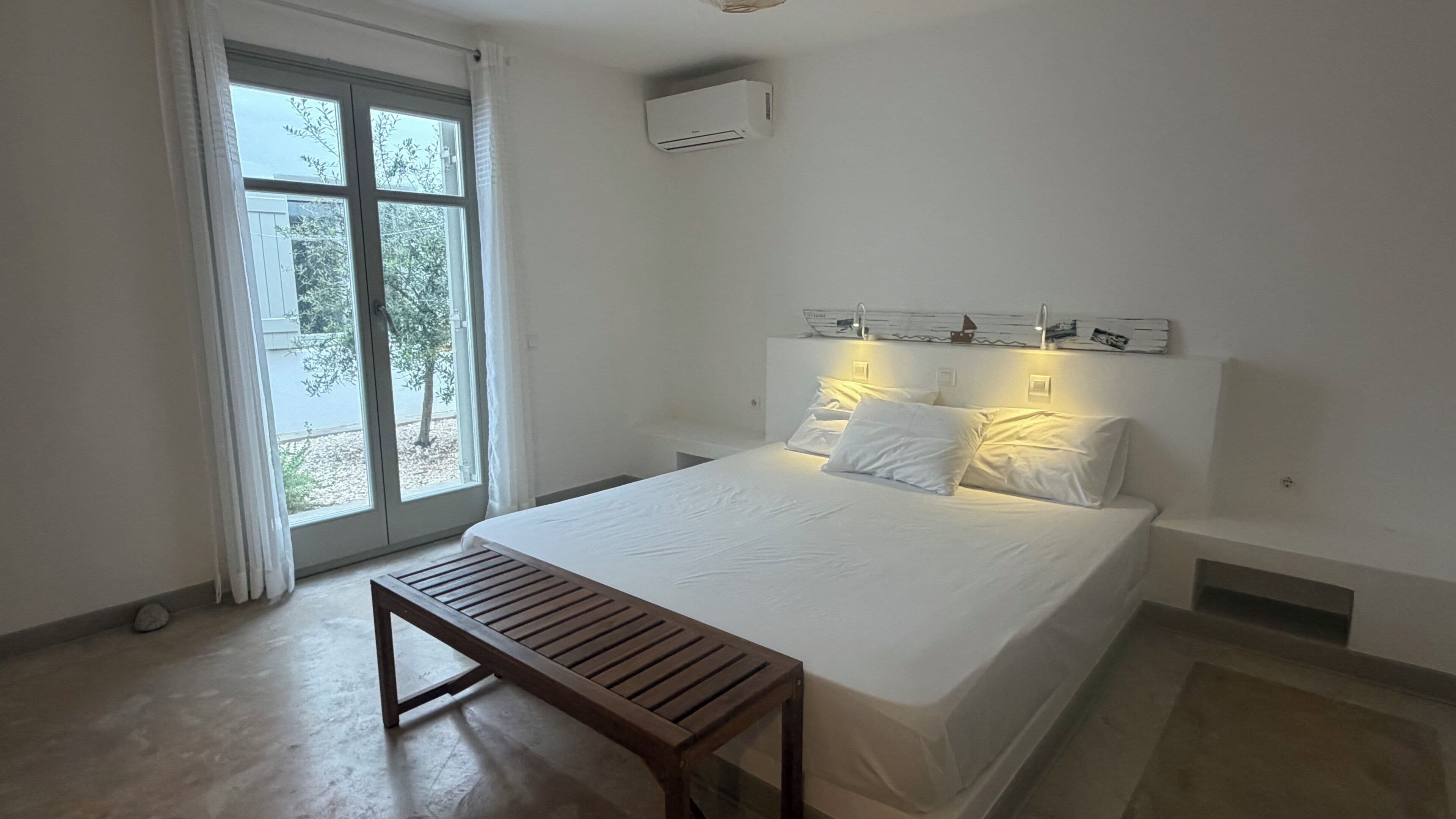 Un dormitorio con una cama, un banco, una ventana y un equipo de aire acondicionado.