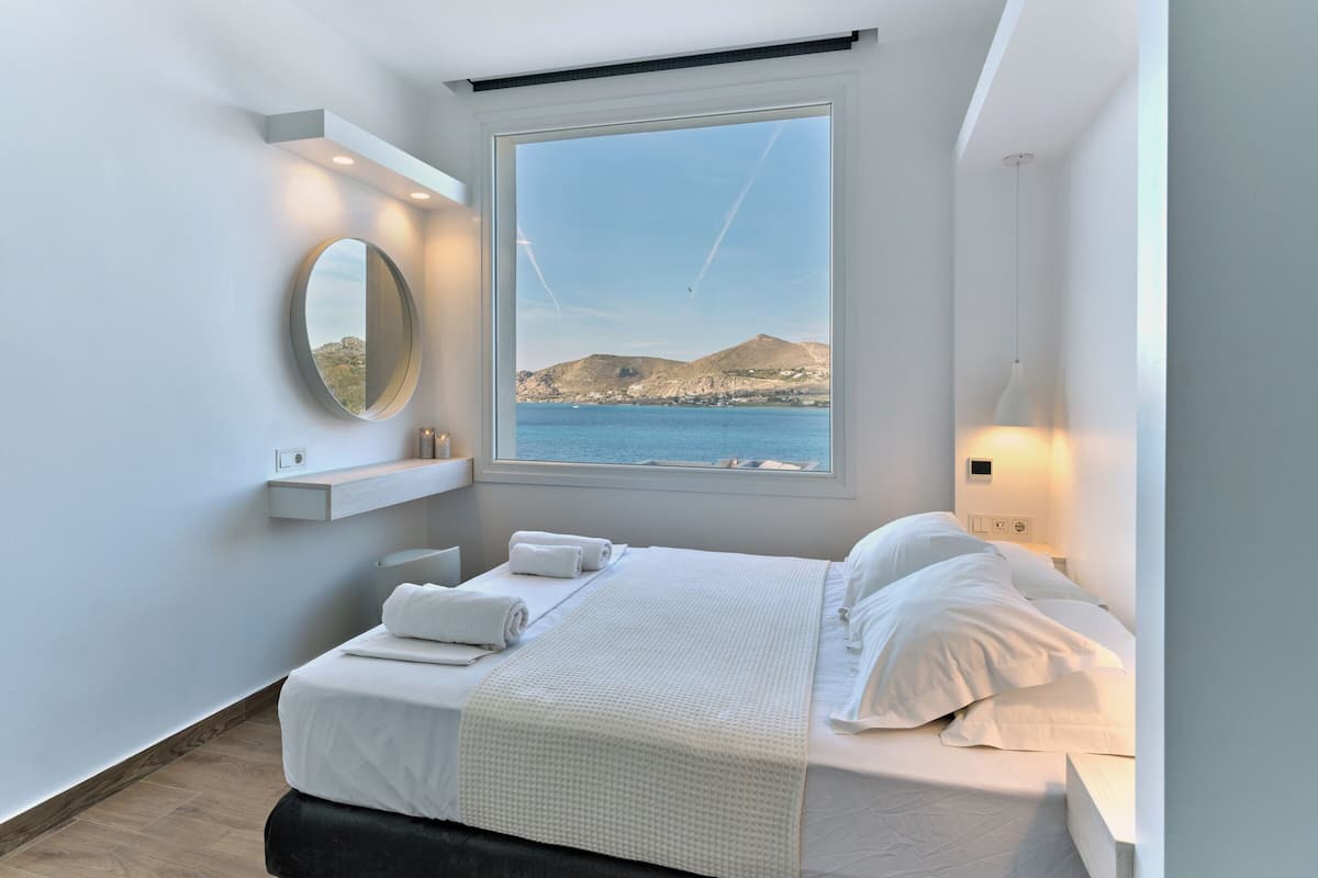 A modern bedroom with a large window, a bed with white linens, a round mirror, and a small shelf with candles.