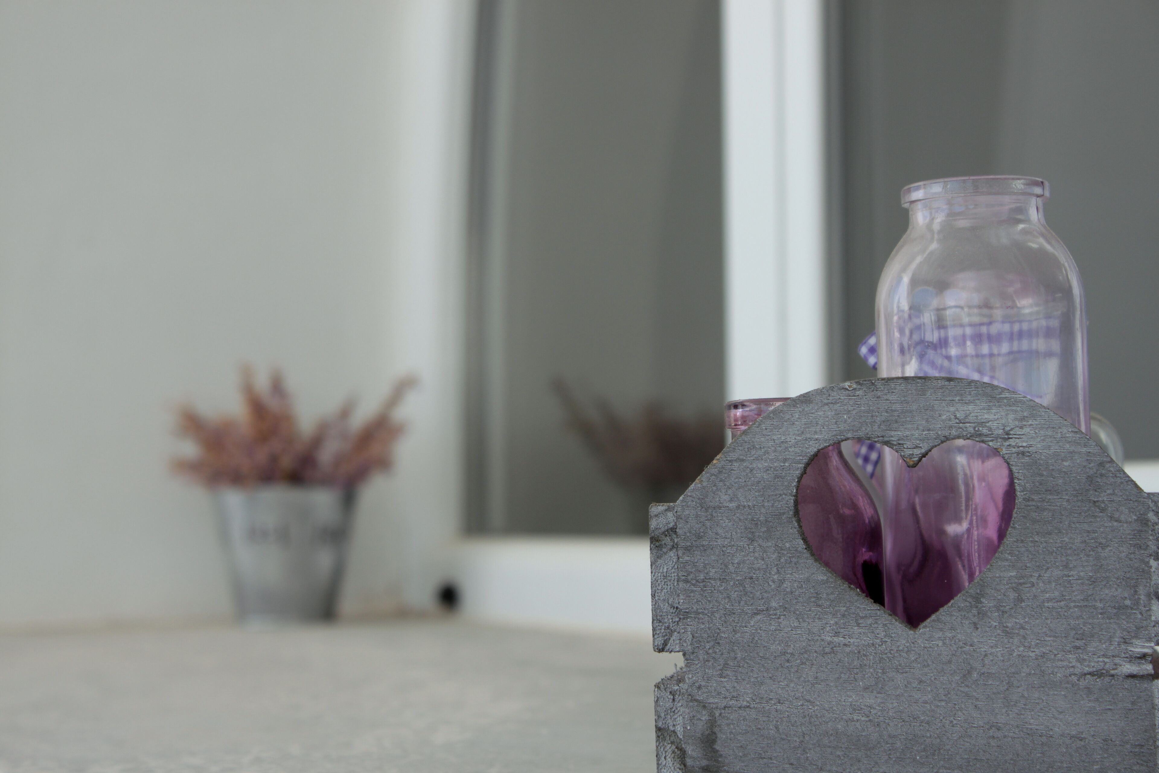 Une enseigne rustique en bois, ornée d’une découpe en forme de cœur, un bocal en verre et un récipient en métal contenant des plantes séchées, le tout sur un fond neutre.