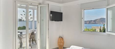 Een moderne slaapkamer met een bed, een televisie, uitzicht op zee en een balkon met een stoel en tafel.
