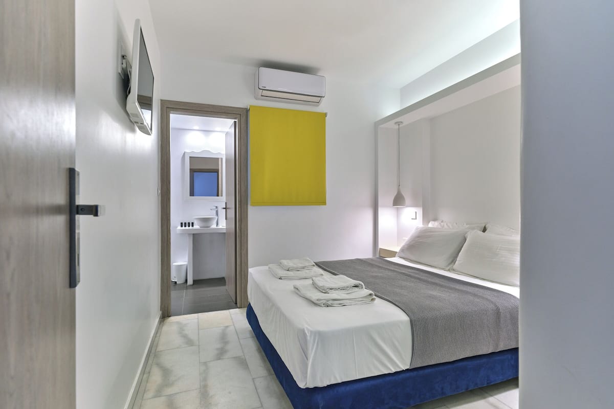 A bedroom with a bed, a bedside table, a nightstand, a mirror, and an air conditioning unit on the wall.