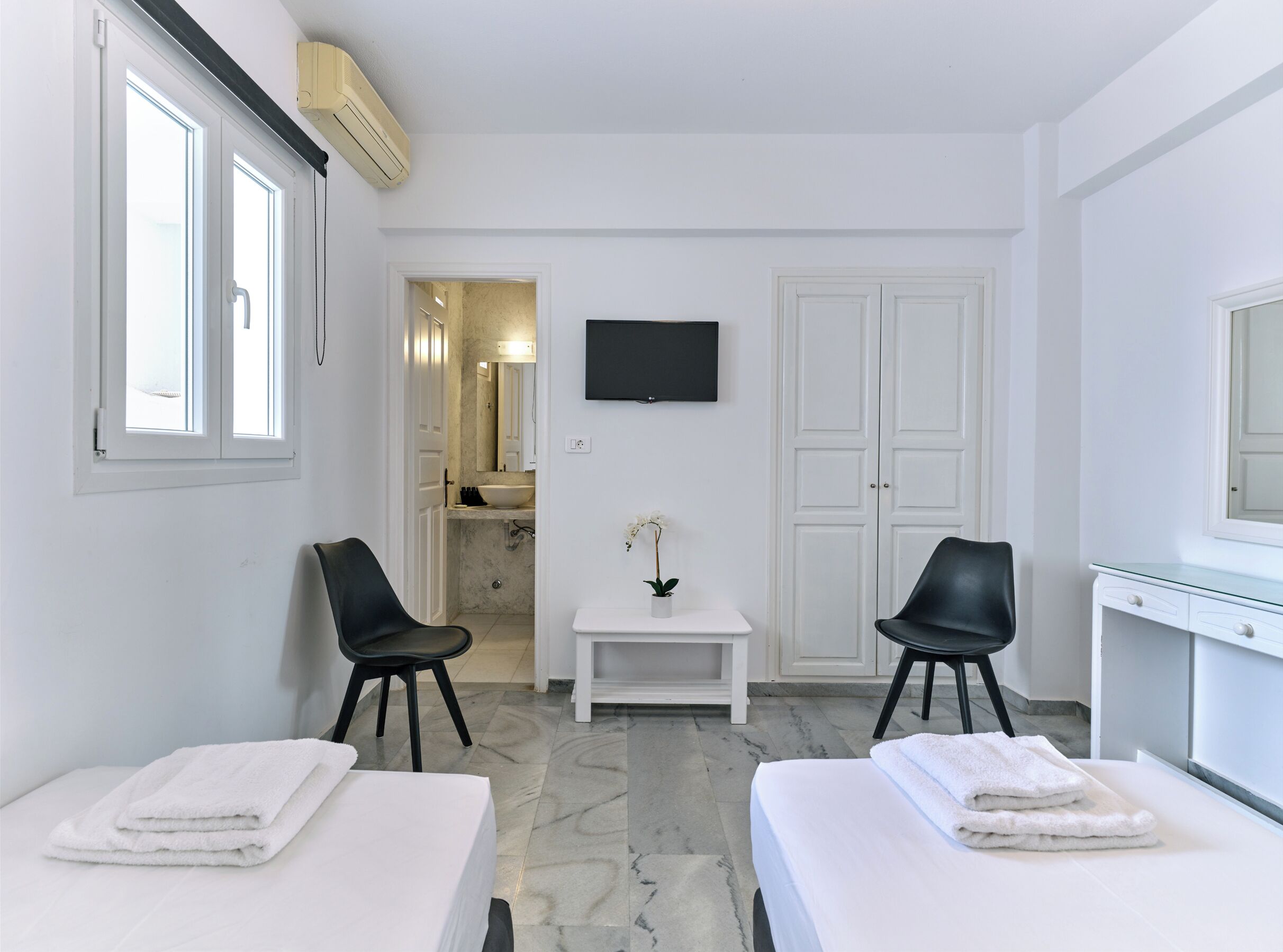 Habitación de hotel con dos camas, un televisor, una mesita con una planta y un baño visible a través de una puerta abierta.