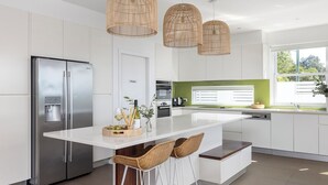 A modern kitchen with a central island, stainless steel appliances, wicker chairs, and a view of the outdoors. - Azure Haven – A Waterfront Luxury Retreat for Families (Corlette)