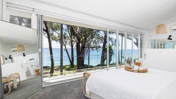A bedroom with a large sliding glass door leading to a balcony with a view of the sea and trees.