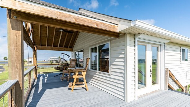 Une terrasse couverte avec une table et des chaises en bois, une vue sur l’océan et un bâtiment blanc doté de grandes fenêtres.