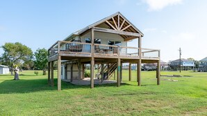 Une maison en bois à deux étages, dotée d’un balcon au niveau supérieur, située dans une zone herbeuse sous un ciel dégagé.