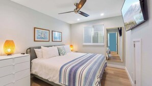 A bedroom with a bed, a ceiling fan, a TV mounted on the wall, and a window with blinds.