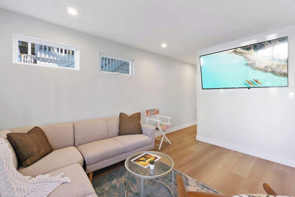 A modern living room with a beige sofa, a glass coffee table, and a flat-screen TV mounted on the wall displaying a scenic coastal view.