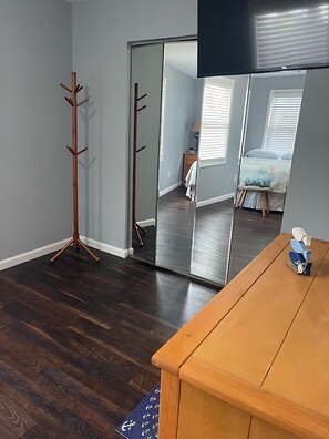 Une pièce comprenant un porte-manteau en bois, une porte de placard avec un miroir, un bureau en bois et un lit recouvert d’une couverture bleue.