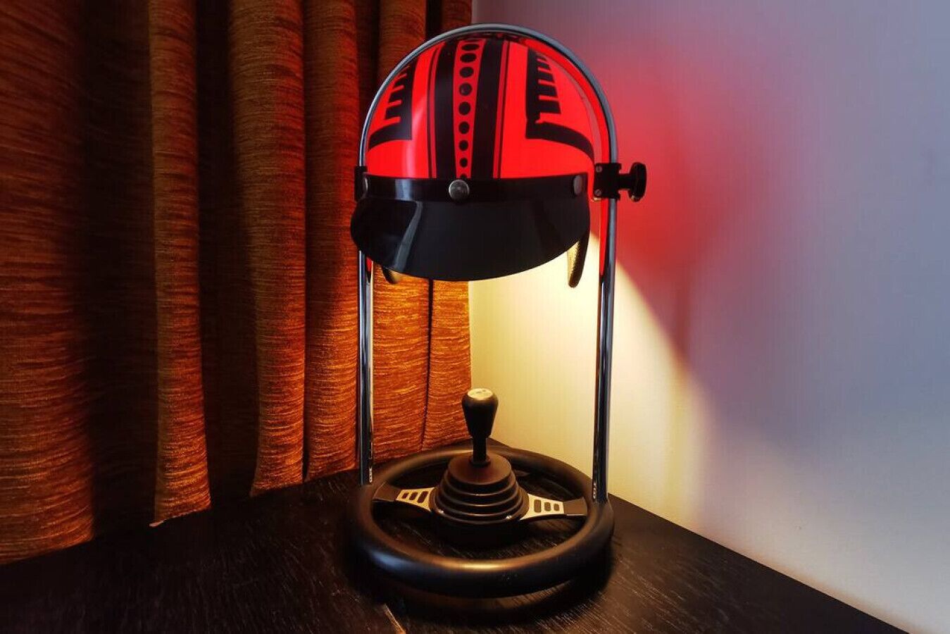Een bureaulamp met een rood en zwart design, een voetstuk met een joystick, en een gordijn op de achtergrond.