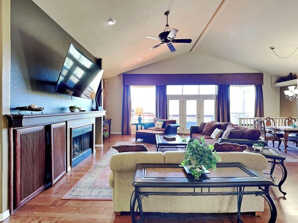 A spacious living room with a fireplace, a television mounted on the wall, a ceiling fan, and a view of the balcony through large windows.
