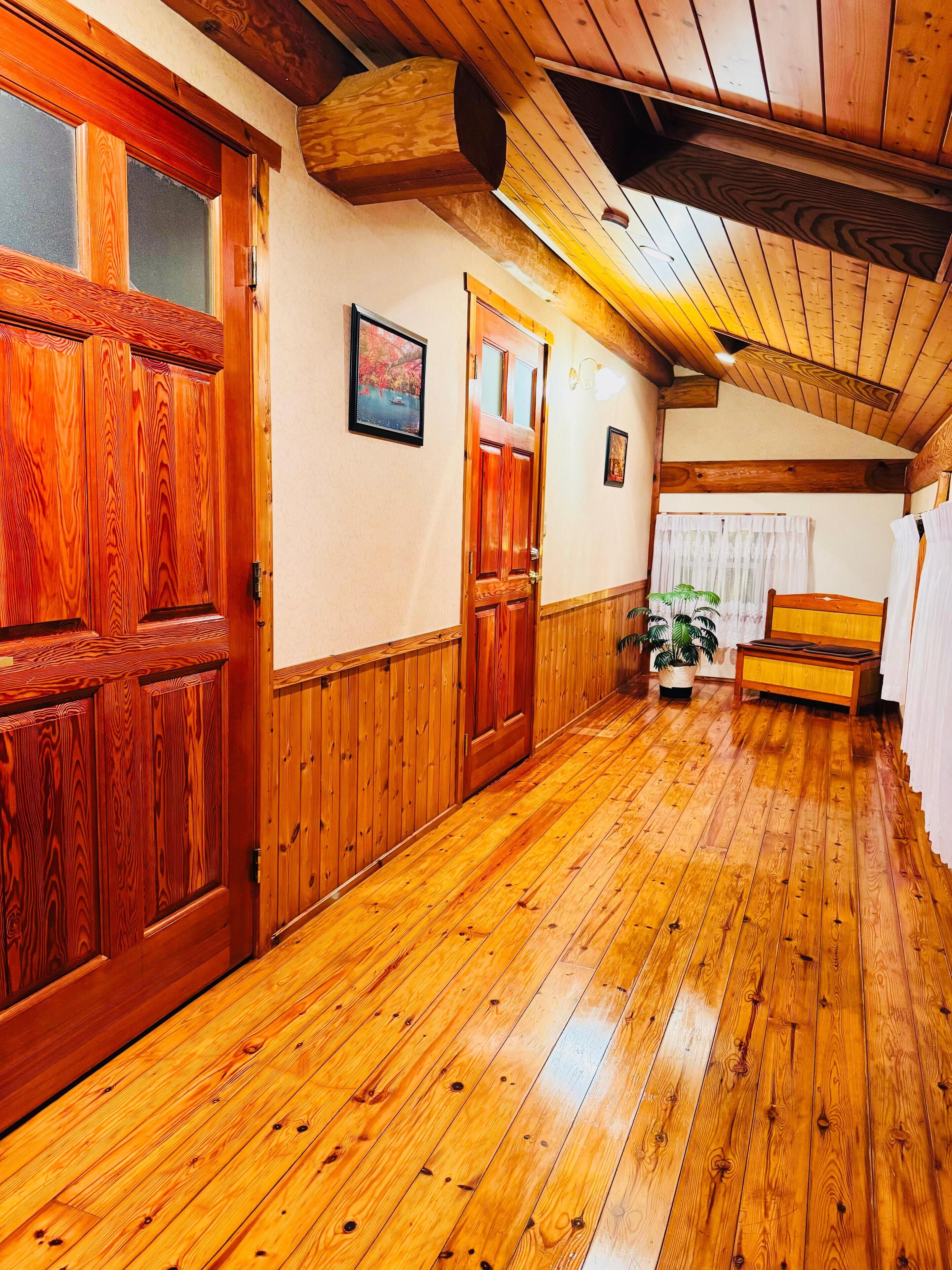 A wooden hallway with a wooden door, a framed picture on the wall, and a bench in the distance.