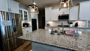 Una cucina moderna con mobili bianchi, elettrodomestici in acciaio inossidabile, piani di lavoro in granito e illuminazione a sospensione.
