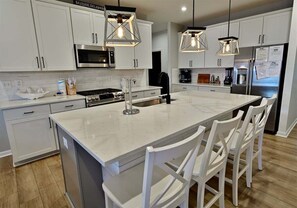 A modern kitchen with white cabinets, a large island, bar stools, stainless steel appliances, and pendant lighting. - New Listing: Bayside Resort: W Fenwick Island Beach House: 5BR 4 (Selbyville)