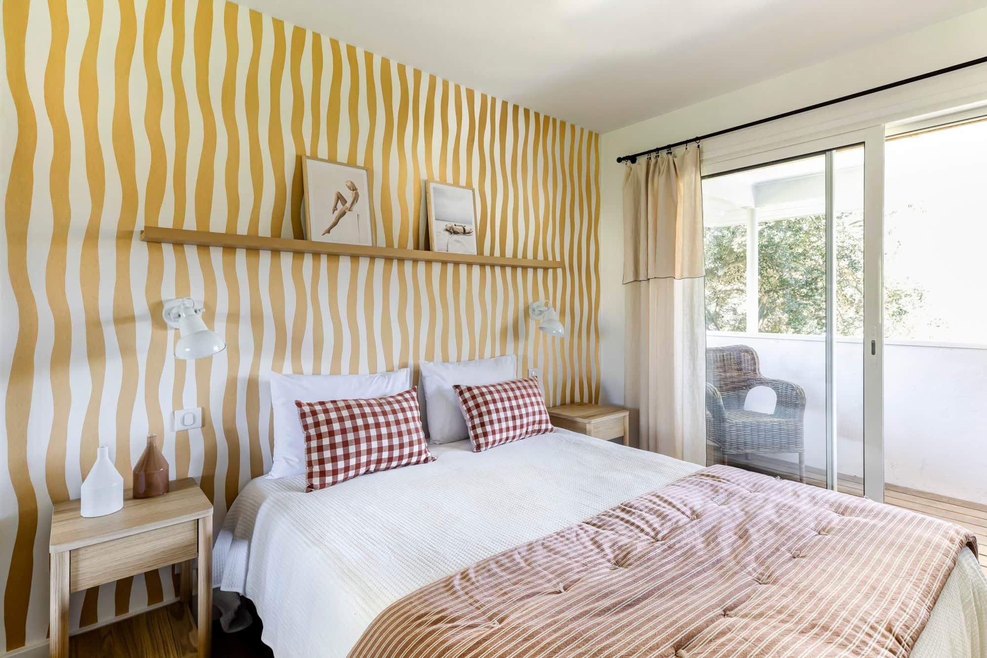 A bedroom with a striped wall, a bed with white bedding, two bedside tables with lamps, and a view of a balcony with a chair.