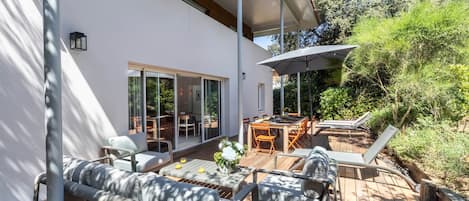 Un patio moderno al aire libre con piso de madera, un sofá, una mesa de comedor y un paraguas grande.