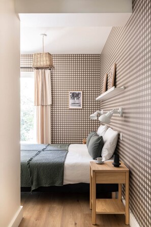 Ein Schlafzimmer mit kariertem Wandpapier, einem Bett mit grüner Decke, einem Nachttisch mit Lampe und einem Fenster mit Vorhängen.