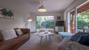 A living room with a brown leather sofa, a white sofa, a coffee table, and a TV on a stand. There is a large window with a view of greenery outside.