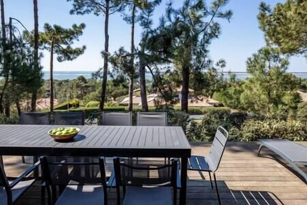 Un área de comedor al aire libre con una mesa y sillas, con vistas a un jardín ajardinado y el mar.