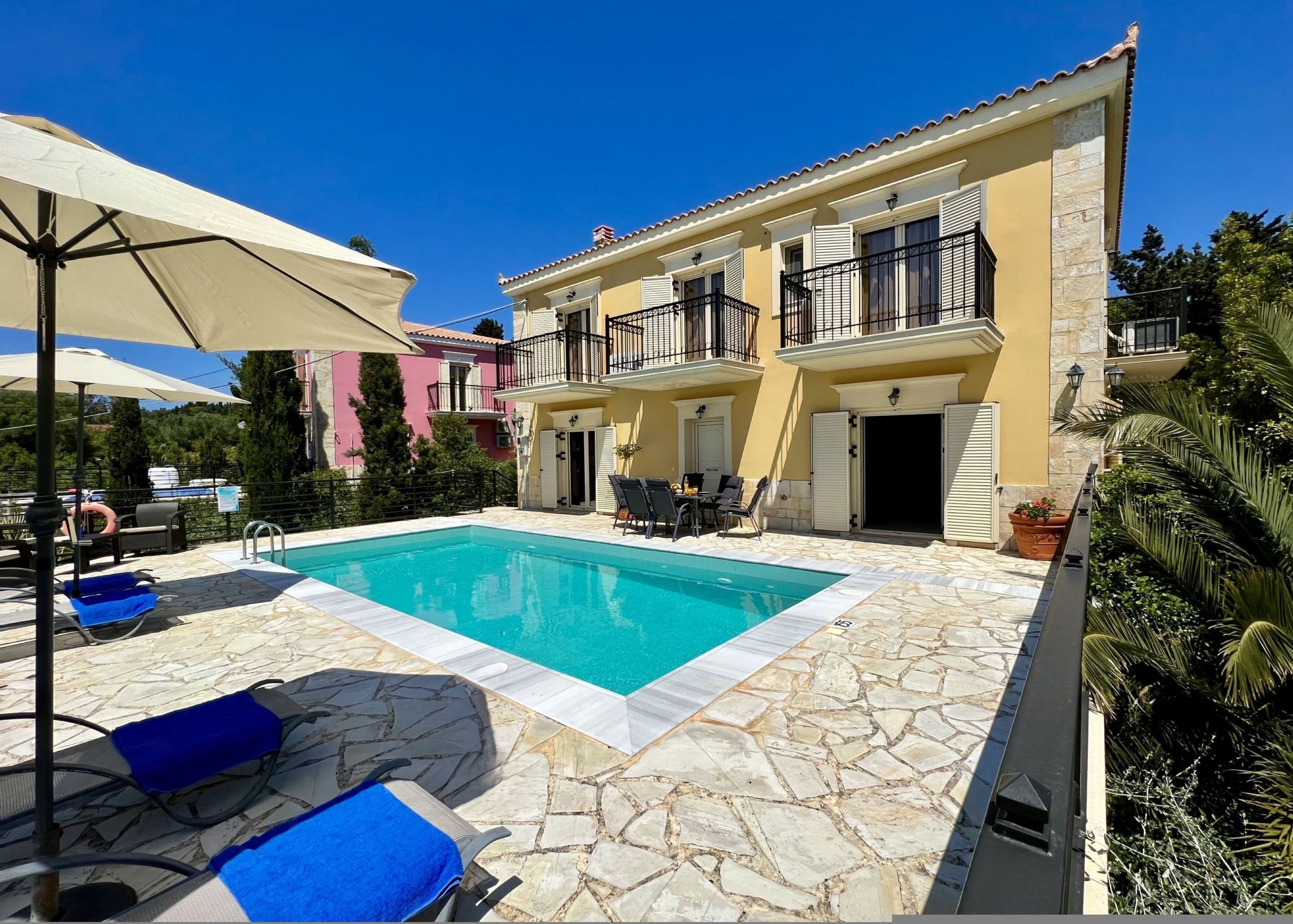 A pool area with lounge chairs and umbrellas, a stone-tiled deck, and a building with balconies and a stone facade.