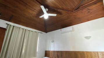 Ein Hotelzimmer mit Holzboden, einem Bett mit weißer Bettwäsche, einem Deckenventilator und einem Fenster mit Vorhängen.