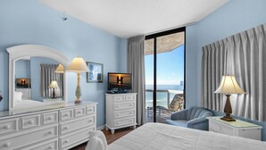 A hotel room with a large bed, a sitting area with a view of the ocean, a television, and a balcony with a chair.