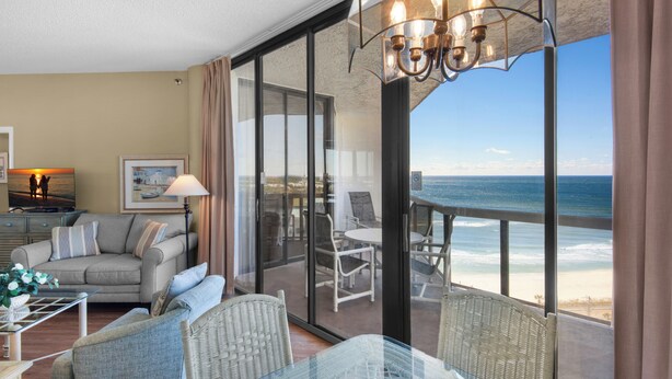 A room with a view of the beach, furnished with a sofa, chairs, and a dining table.