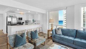 A modern living room with a blue sofa, a dining area with a kitchen, and a large window offering a view of the ocean.
