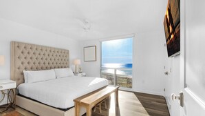 A bedroom with a large bed, a wooden bench, a nightstand, and a view of the ocean.