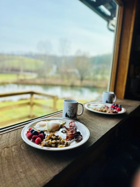 En morgenmadsopsætning med tallerkener med mad og en kop på et træbord ved et vindue med udsigt over vand.