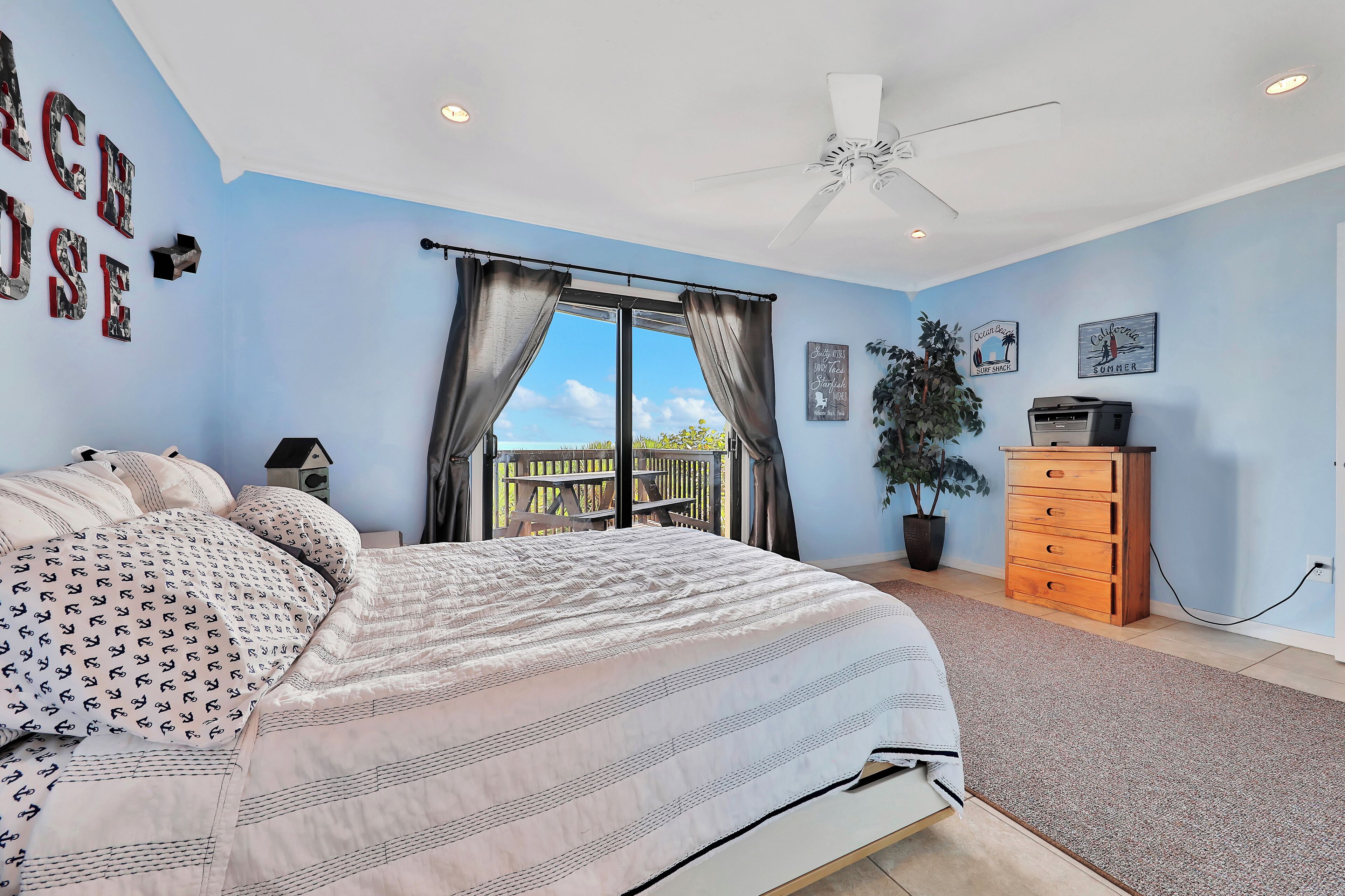 A bedroom with a bed, a ceiling fan, a dresser, a potted plant, and a view of the outdoors.