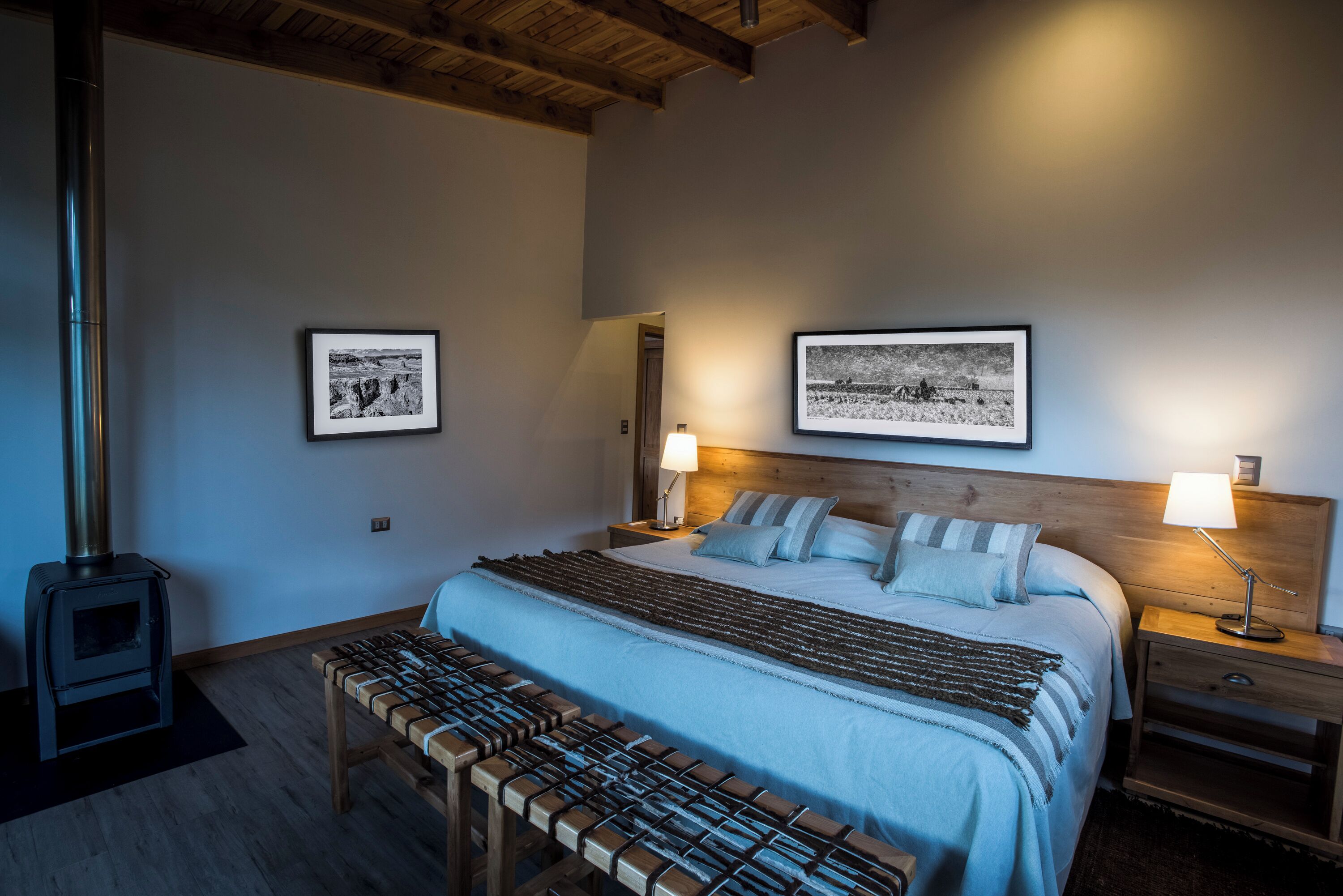 Quarto com uma cama grande, duas mesas de cabeceira, um banco de madeira, aquecedor fixado na parede e duas imagens emolduradas.