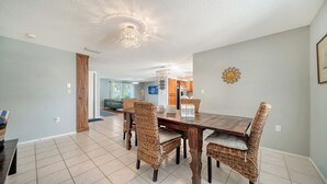 A dining area with a wooden table and wicker chairs, a kitchen in the background, and a ceiling light fixture.